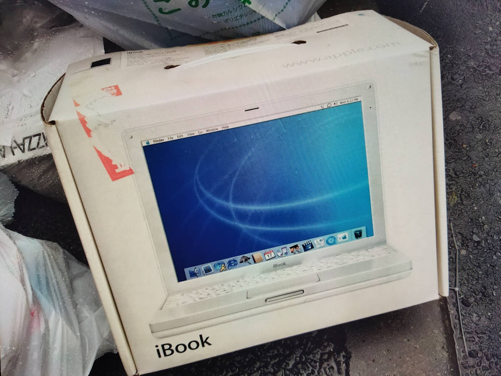 In a quiet corner of the street, an iBook G3 Snow box rests among trashed items, a poignant reminder of the ever-evolving nature of technology and the cherished memories of its early days.